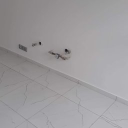 Marcin Wadelski - Przygotowane podejścia wodno-kanalizacyjne w nowej łazience z białymi płytkami imitującymi marmur na podłodze i gładzią na ścianach.