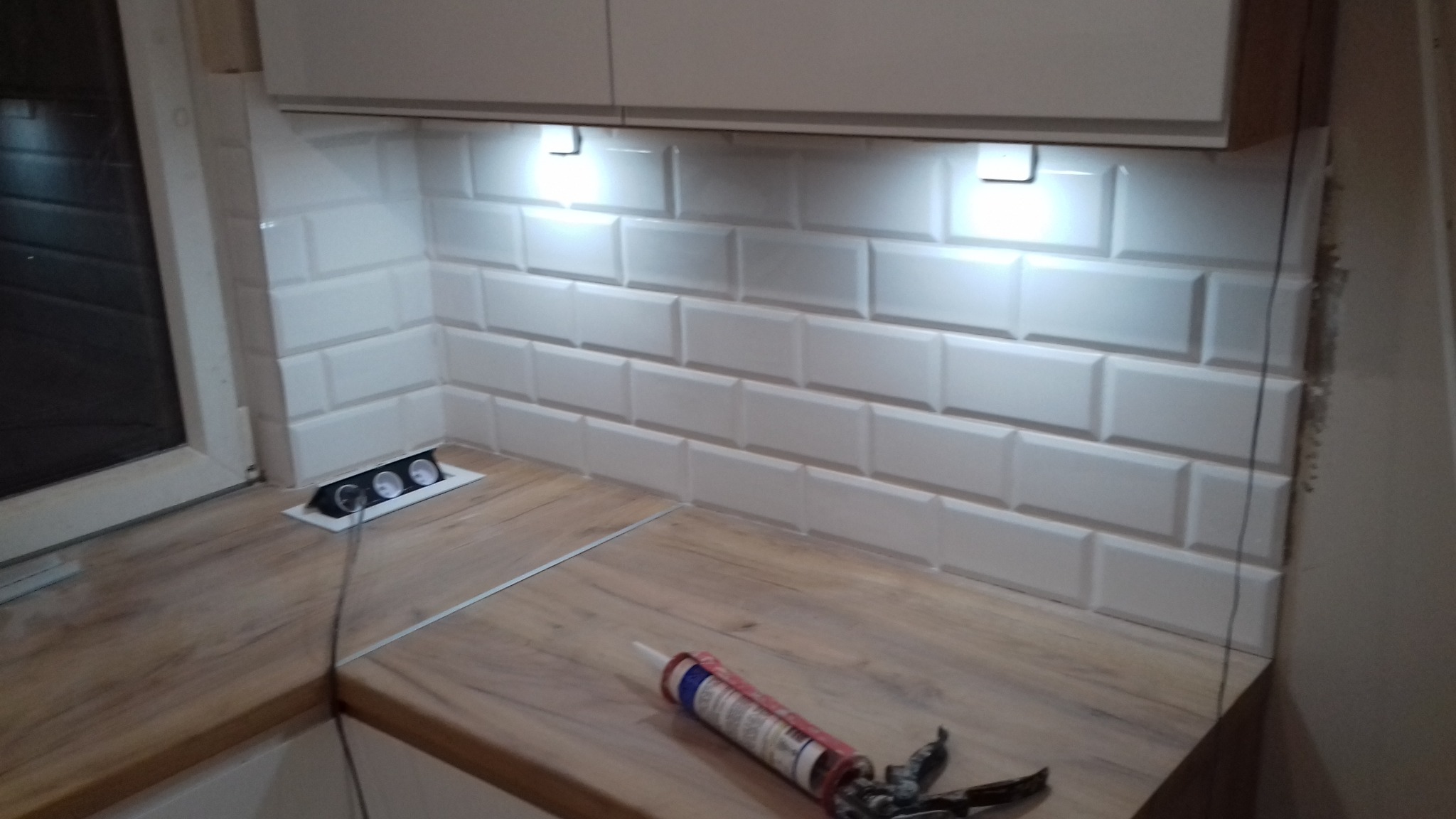 Białe, prostokątne płytki ułożone na ścianie w kuchni pod szafkami, oświetlone punktowymi lampkami, z widocznym gniazdkiem elektrycznym w blacie oraz tubką silikonu i pistoletem do silikonu na blacie.