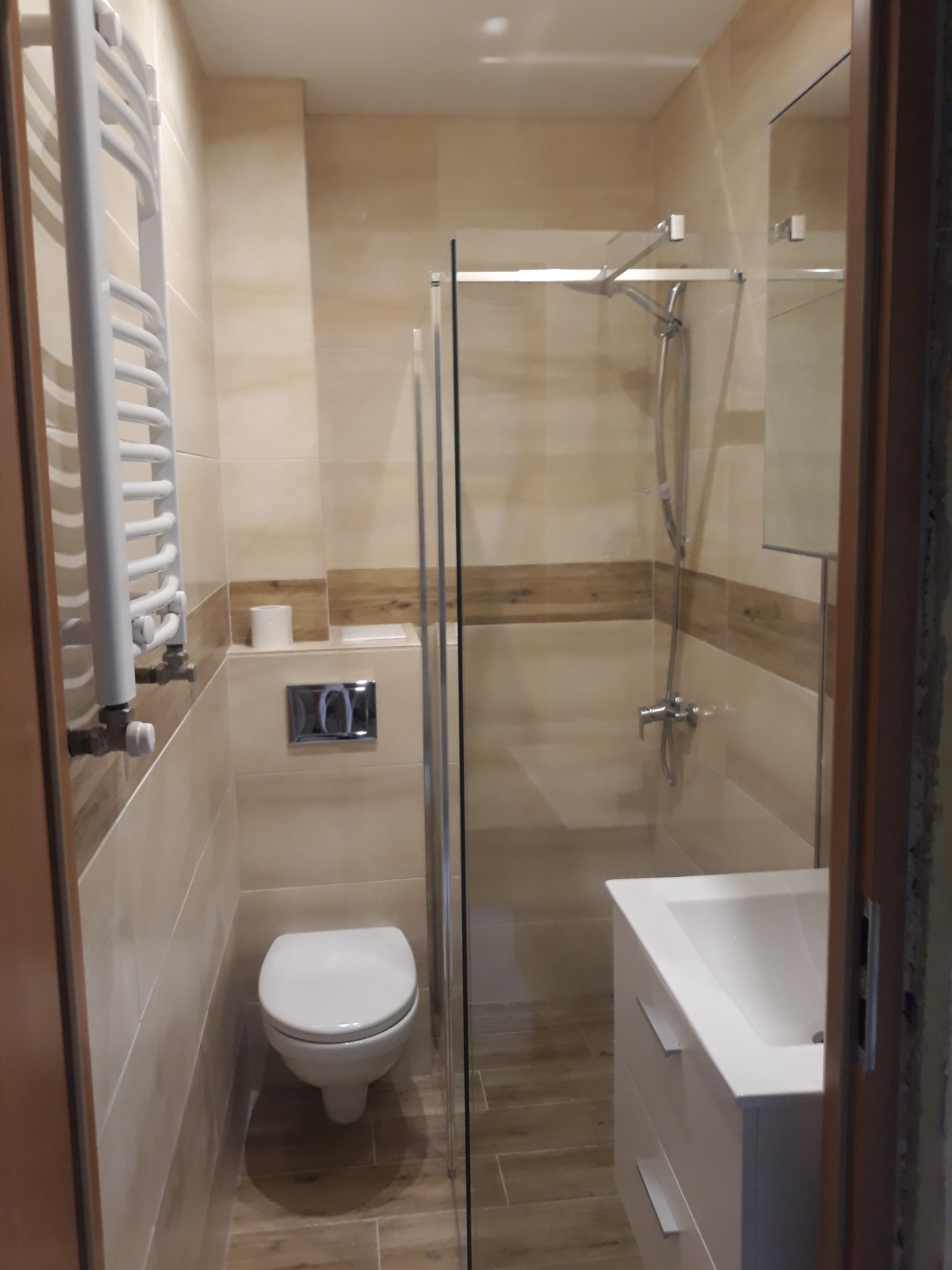 Nowoczesna łazienka z kabiną prysznicową za szklaną ścianką, wiszącą toaletą z chromowanym przyciskiem spłukiwania, białą umywalką z szafką i grzejnikiem drabinkowym w jasnych beżowych płytkach...