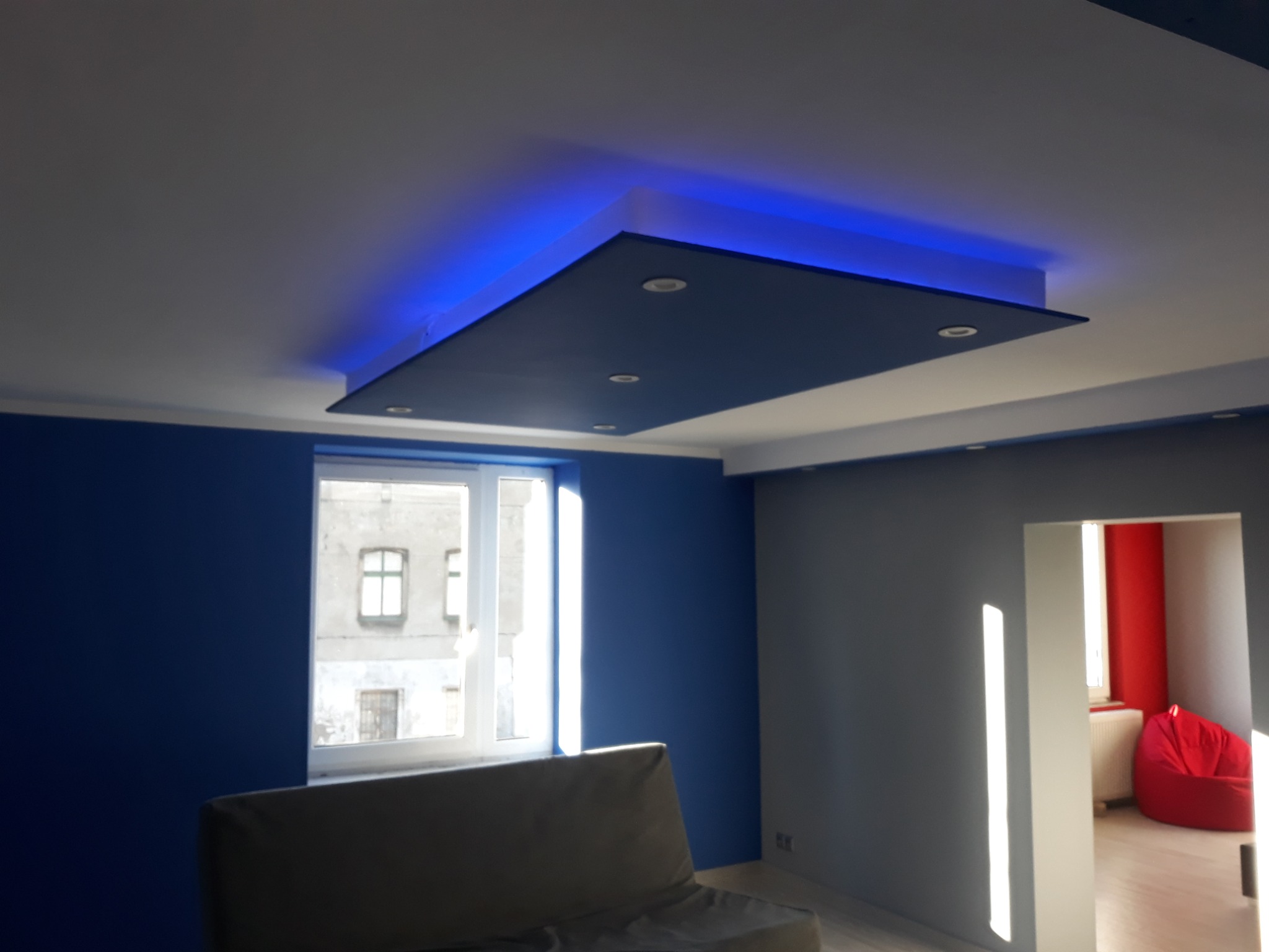 Nowoczesny pokój z podwieszanym, podświetlanym na niebiesko sufitem z wbudowanymi punktami świetlnymi, niebieskimi ścianami i widokiem na budynek z okna