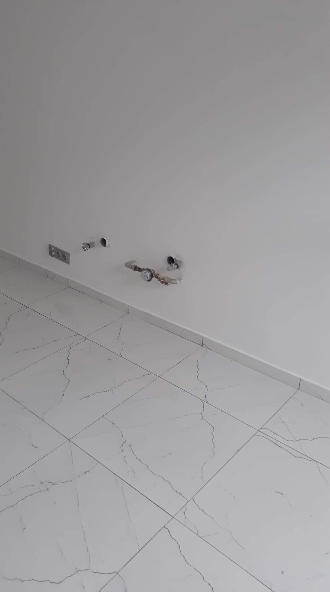 Przygotowane podejścia wodno-kanalizacyjne w nowej łazience z białymi płytkami imitującymi marmur na podłodze i gładzią na ścianach.