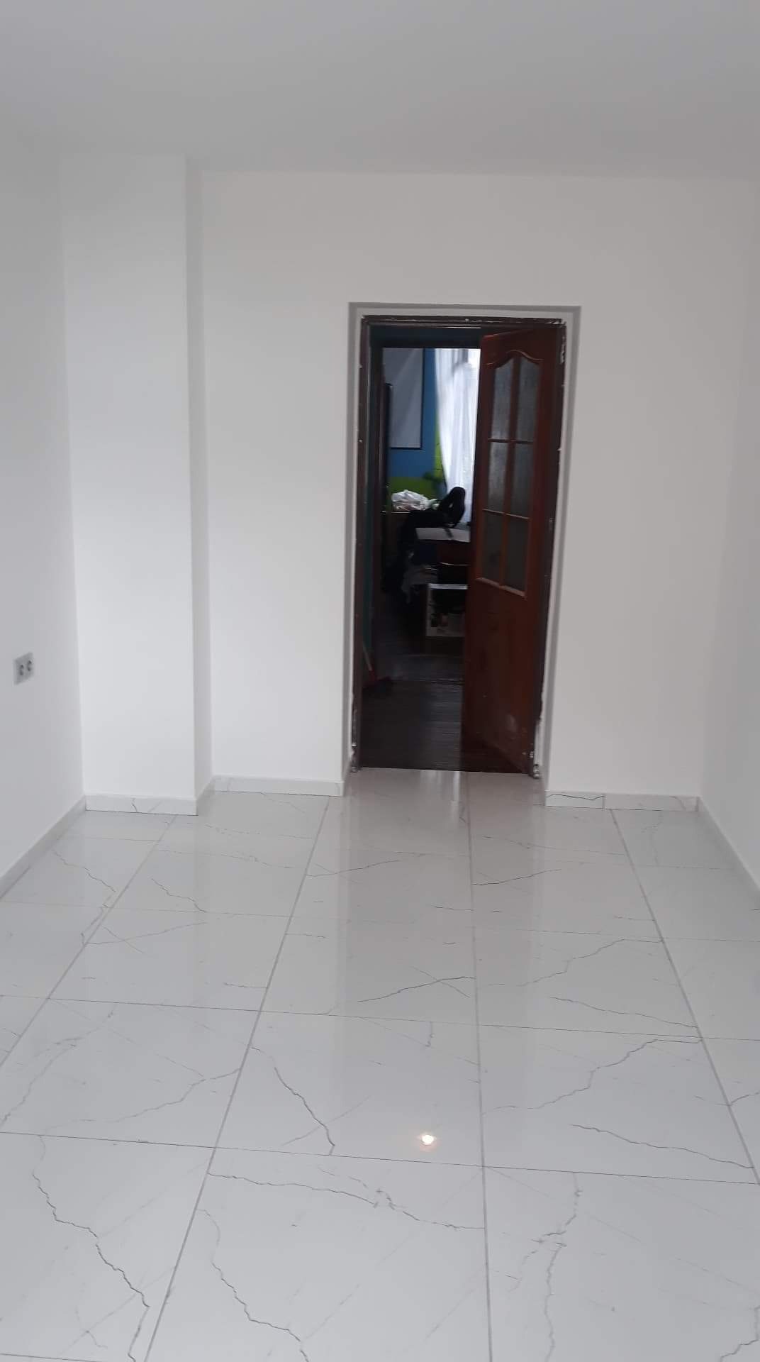 Wnętrze pokoju po remoncie: białe ściany, białe płytki podłogowe z szarym wzorem żyłek, otwarte drewniane drzwi do sąsiedniego pokoju.