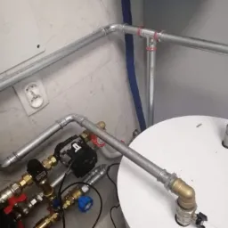 Szare rury instalacji hydraulicznej z licznymi zaworami i pompą obiegową, zamontowane w rogu pomieszczenia z marmurową ścianą i fragmentem niebieskiego zbiornika.