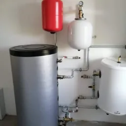 Kompletna instalacja grzewcza z zbiornikiem akumulacyjnym, czerwonym i białym zbiornikiem wyrównawczym, oraz rurami instalacyjnymi z zaworami i manometrami, umieszczona przy białej ścianie.