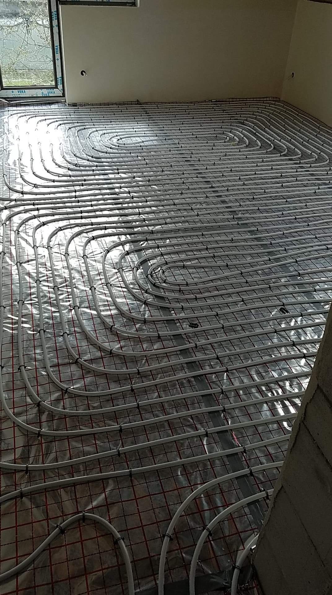 Rozłożona instalacja ogrzewania podłogowego z rur na folii z siatką, widok z góry, surowy stan wykończenia wnętrza.