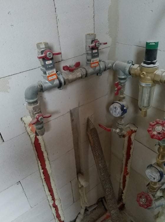 Szare rury instalacji wodnej z czerwonymi zaworami, liczniki wody i filtr, zamontowane na ścianie z betonu komórkowego z widoczną pianką montażową w szczelinach.