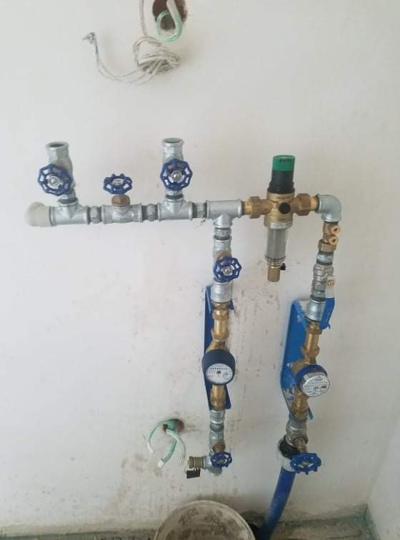 Kompleksowa instalacja wodociągowa z licznymi zaworami i filtrem, widoczne dwa wodomierze zamontowane na niebieskich wspornikach, ściana w trakcie wykończenia.