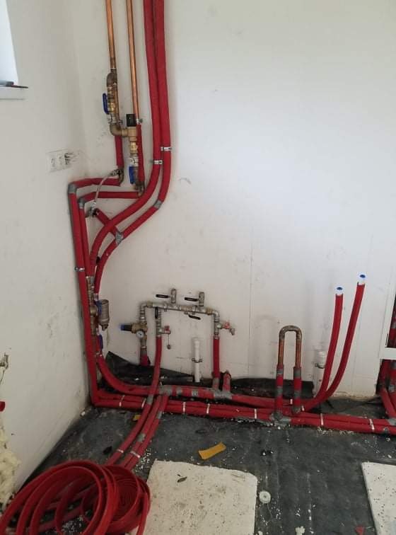 Kompleksowa instalacja hydrauliczna w trakcie montażu: czerwone rury PEX prowadzone wzdłuż ściany i podłogi, widoczne zawory, złączki i fragment zwiniętej rury, surowe wykończenie wnętrza.