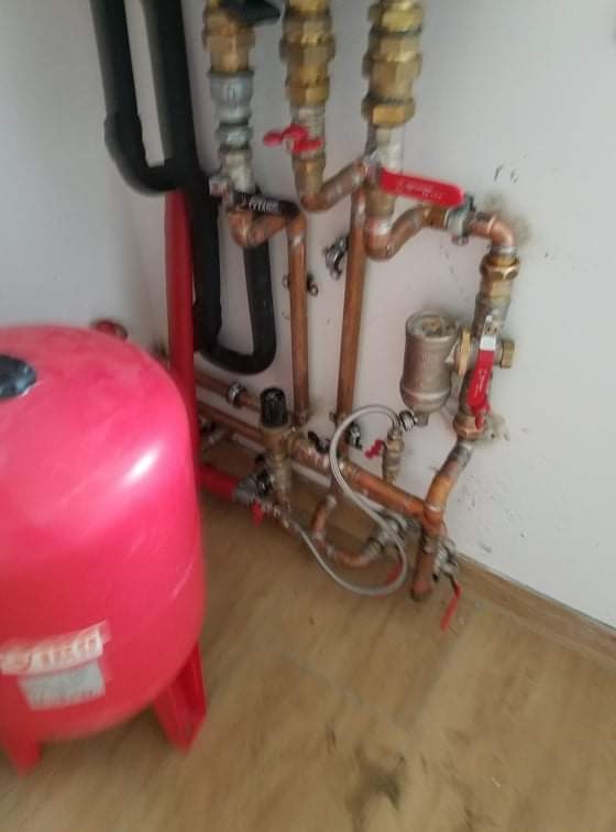 Instalacja hydrauliczna z czerwonym zbiornikiem wyrównawczym i miedzianymi rurami w Trawnikach, widoczne zawory i filtr siatkowy.