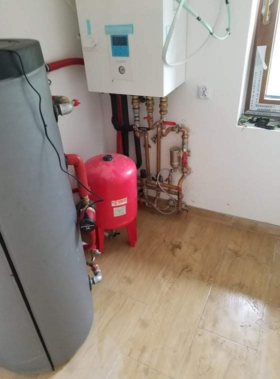 Kotłownia z szarym zasobnikiem ciepłej wody, czerwonym naczyniem wzbiorczym IBO, białym kotłem gazowym i miedzianą instalacją hydrauliczną na tle beżowych płytek podłogowych.