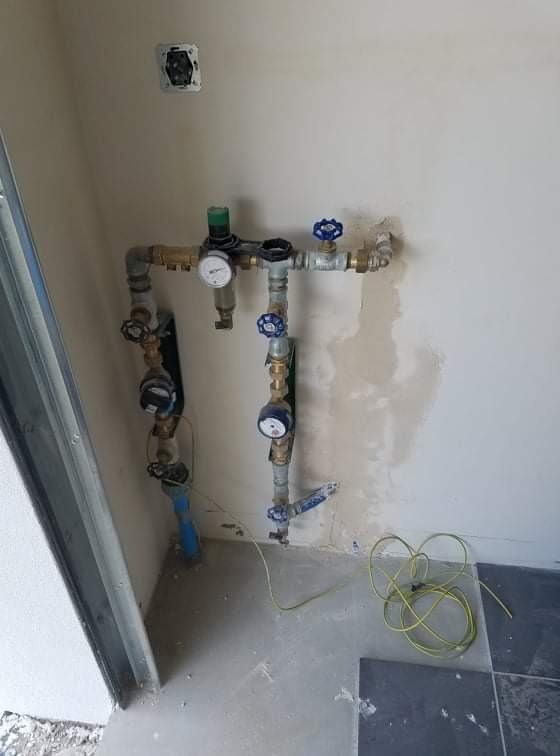 Zestaw rur wodociągowych z zaworami, manometrami i filtrem, zamontowany na surowej ścianie w trakcie prac wykończeniowych. Widoczne przewody elektryczne na podłodze.