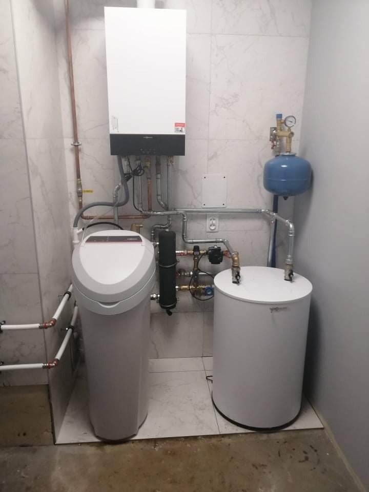 Kompaktowa kotłownia domowa z białym kotłem wiszącym, zmiękczaczem wody i zbiornikiem wyrównawczym, instalacja w pomieszczeniu wykończonym marmurowymi płytkami.