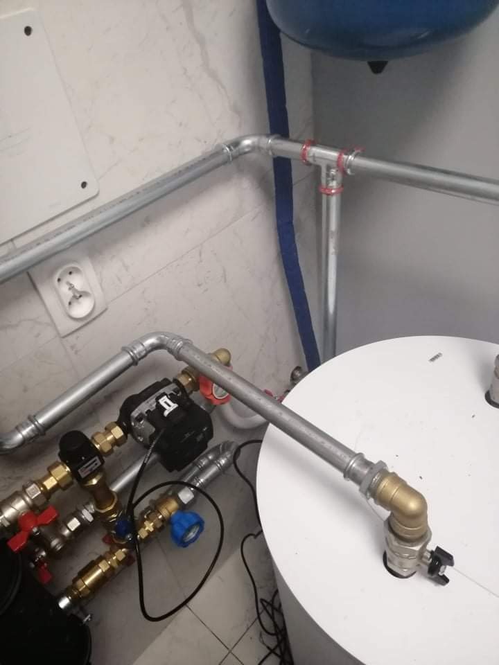 Szare rury instalacji hydraulicznej z licznymi zaworami i pompą obiegową, zamontowane w rogu pomieszczenia z marmurową ścianą i fragmentem niebieskiego zbiornika.