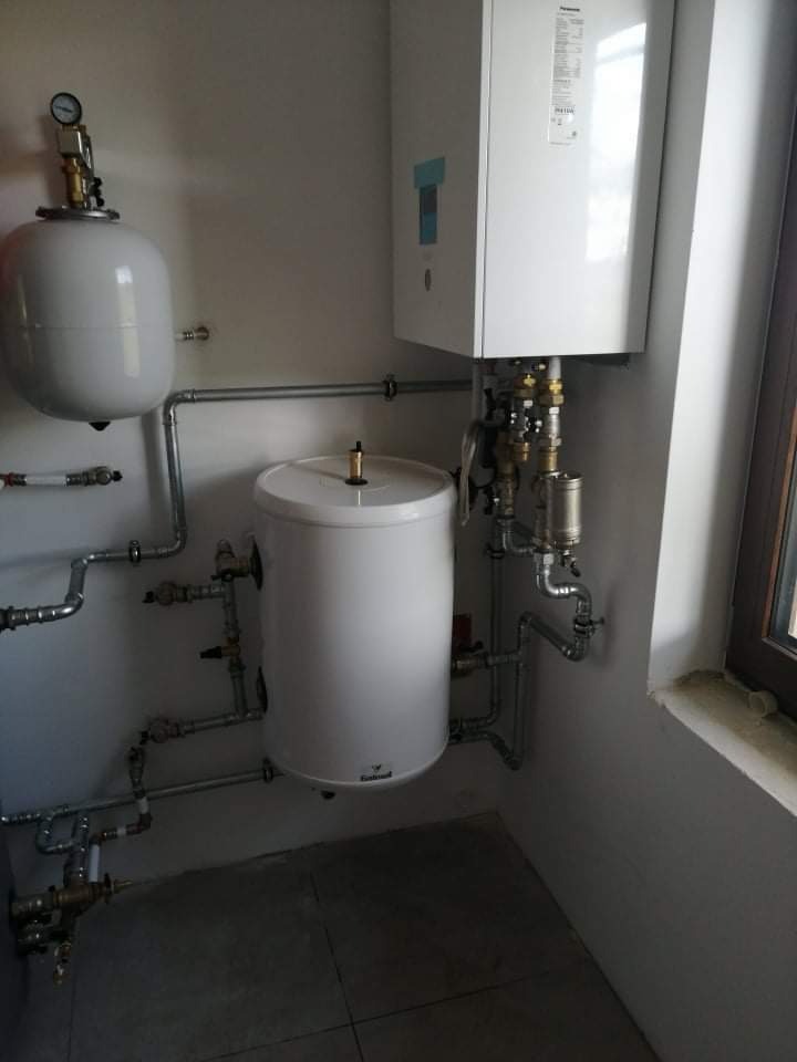 Zestaw instalacji centralnego ogrzewania z białym kotłem i zbiornikami, widoczne rury miedziane i armatura, zamontowane przy ścianie w pomieszczeniu z oknem.