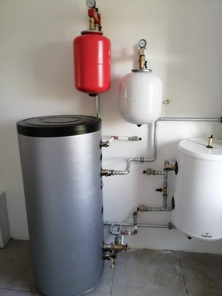 Kompletna instalacja grzewcza z zbiornikiem akumulacyjnym, czerwonym i białym zbiornikiem wyrównawczym, oraz rurami instalacyjnymi z zaworami i manometrami, umieszczona przy białej ścianie.