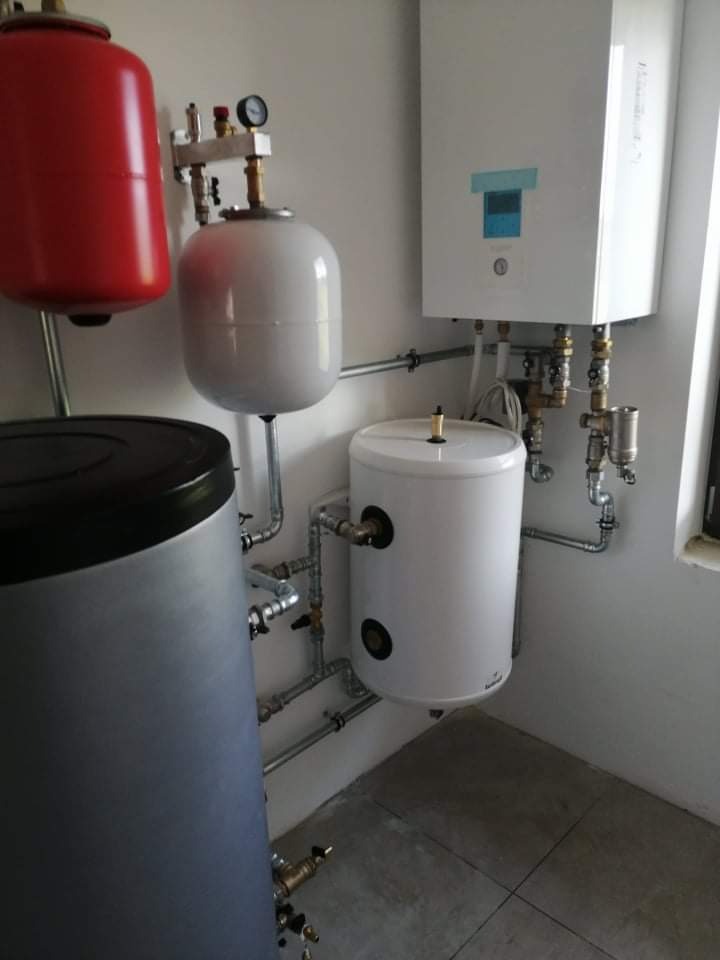 Kompleksowa instalacja centralnego ogrzewania z zasobnikami wody o różnych pojemnościach i kolorach (czerwony, biały, szary), z licznymi rurami i zaworami, zamontowana w pomieszczeniu z szarą podłogą.