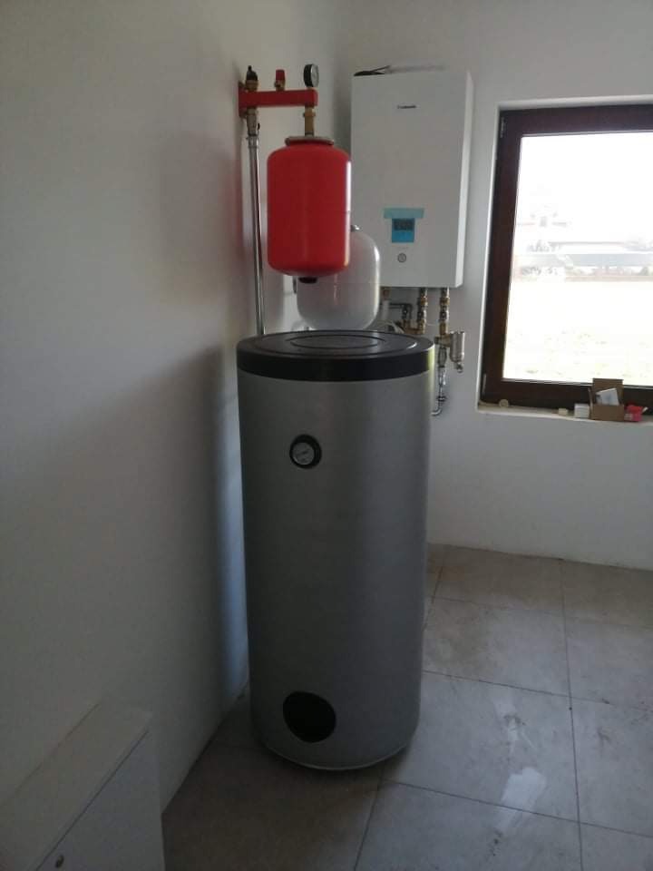 Nowoczesny, srebrny bojler z czerwonym zbiornikiem wyrównawczym i białym piecem gazowym zamontowany na ścianie w jasnym pomieszczeniu z oknem.