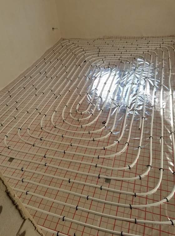 Białe rury ogrzewania podłogowego, ułożone w pętle na izolacji z siatką w czerwone kwadraty, zabezpieczone czarnymi klipsami, widok z góry w surowym wnętrzu.