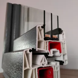 Przekrój poprzeczny ramy okiennej z trzema szybami, ukazujący wielokomorową konstrukcję profilu z uszczelkami i elementami izolacyjnymi, z szarą okleiną zewnętrzną imitującą strukturę drewna.