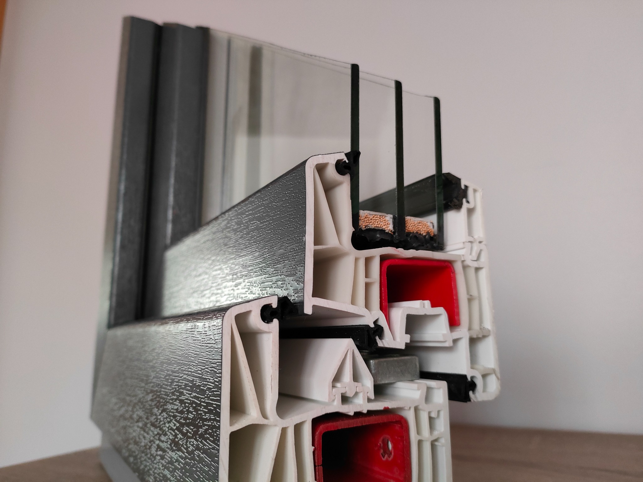 Przekrój poprzeczny ramy okiennej z trzema szybami, ukazujący wielokomorową konstrukcję profilu z uszczelkami i elementami izolacyjnymi, z szarą okleiną zewnętrzną imitującą strukturę drewna.