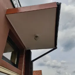 Zewnętrzny podbitka dachowa w kolorze brązowym, z zamontowanym oświetleniem punktowym, widoczna z dołu na tle pochmurnego nieba.