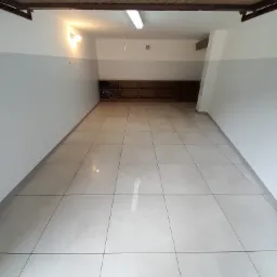 Puste pomieszczenie z wykafelkowaną podłogą, białe ściany z szarym pasem, oświetlone lampą sufitową i żarówkami, z drewnianą wnęką na końcu pomieszczenia.