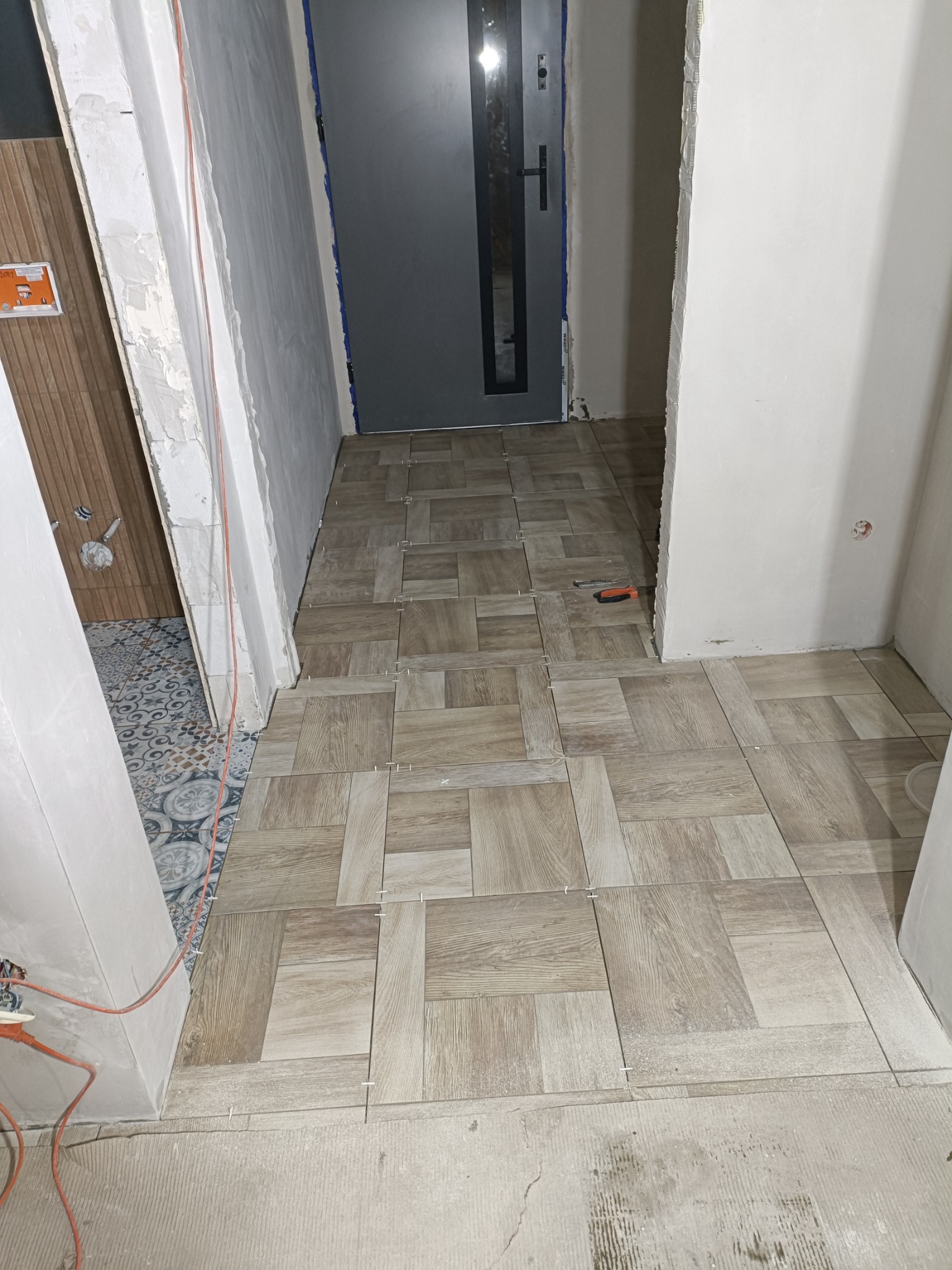 Układanie jasnych, prostokątnych płytek imitujących drewno na podłodze w korytarzu, widoczne krzyżyki dystansowe, niedokończone ściany i drzwi wejściowe w tle.