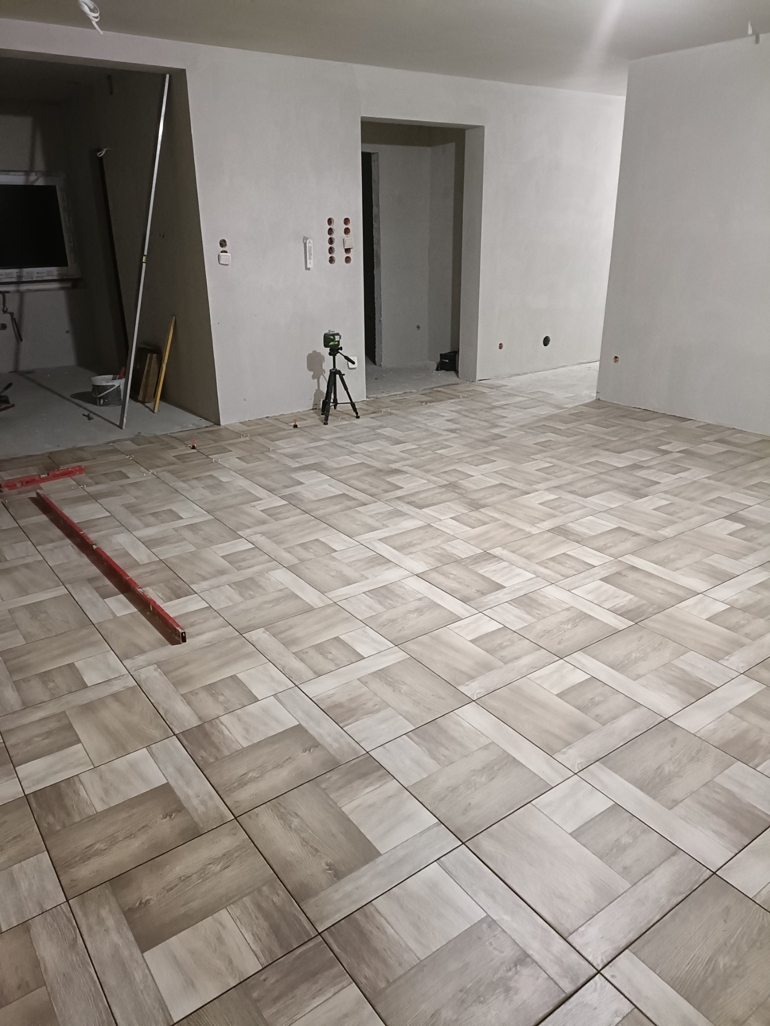 Układanie płytek podłogowych w jodełkę z użyciem poziomicy i lasera krzyżowego w surowym wnętrzu.