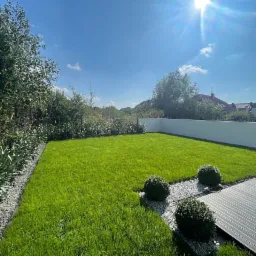 Świeżo założony trawnik z rolki, obrzeża z szarych kamieni, kuliste krzewy ozdobne i nowoczesny taras z desek kompozytowych w słoneczny dzień.