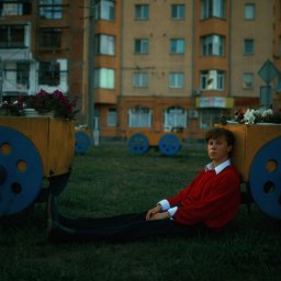 Natalia Czyż - Młody mężczyzna w czerwonym swetrze oparty o dekoracyjny wózek z kwiatami na tle bloku mieszkalnego.