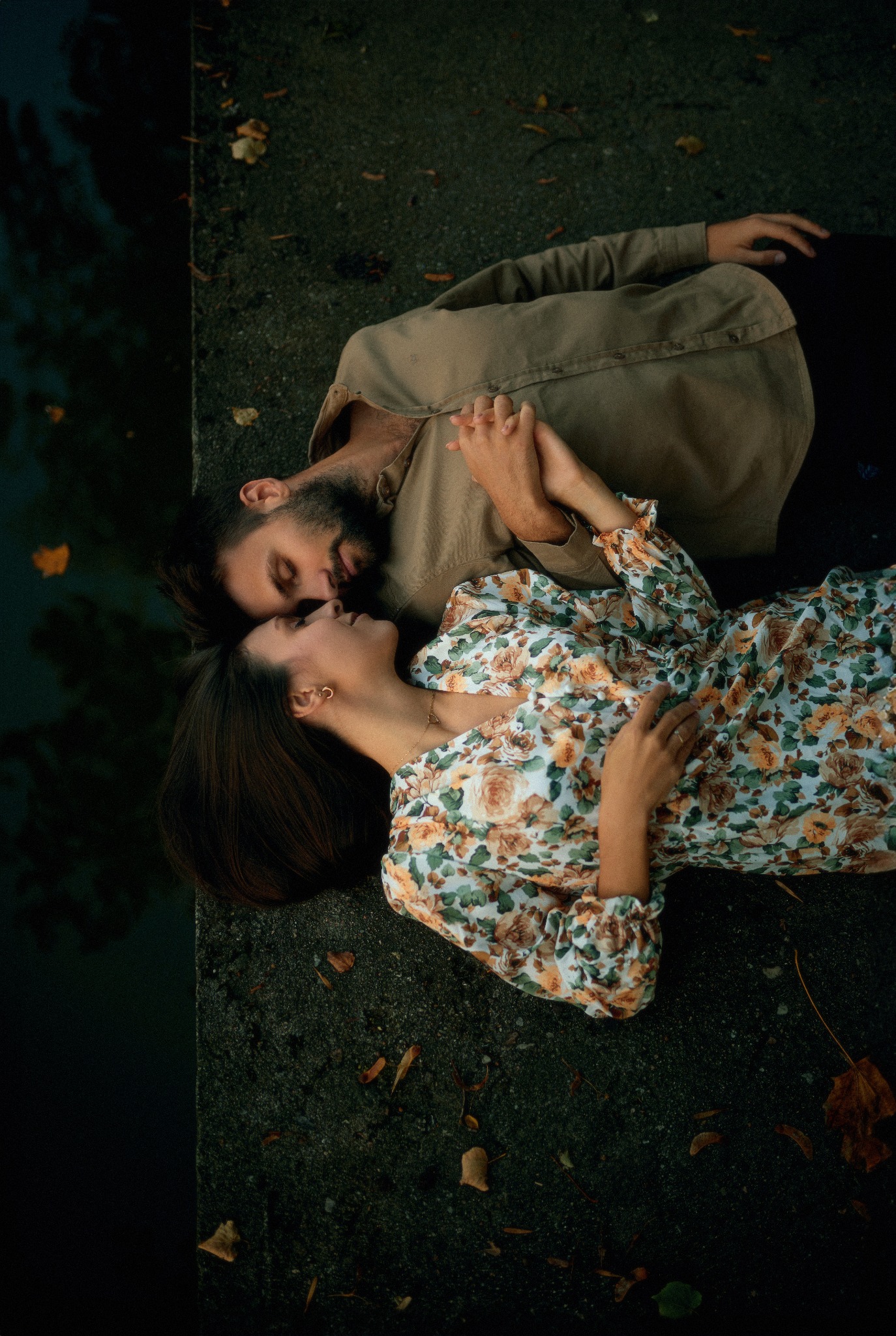 Para zakochanych leży na betonowym murku przy zbiorniku wodnym, trzymając się za ręce; kobieta w kwiecistej sukience, mężczyzna w beżowej koszuli, jesienne liście rozrzucone wokół.