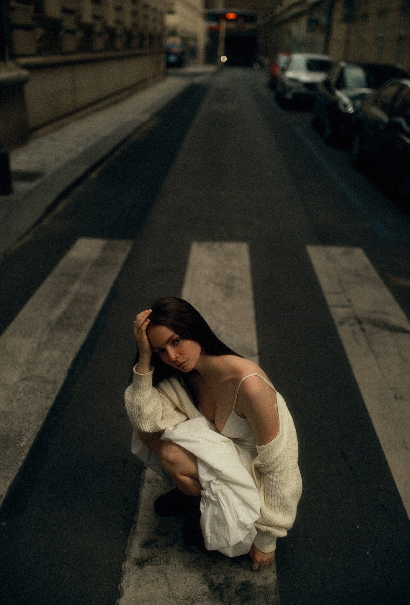 Kobieta w jasnej sukience i kremowym kardiganie kuca na przejściu dla pieszych na tle miejskiej ulicy, zamyślona poza i delikatne oświetlenie.