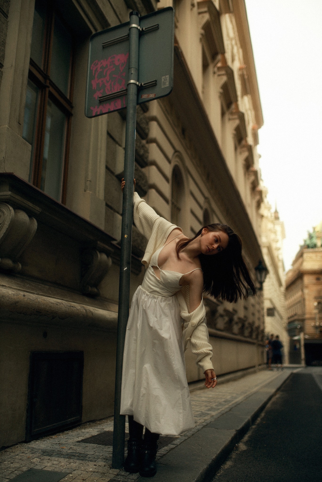 Kobieta w białej sukience i kremowym kardiganie opiera się o słup z drogowskazem w miejskiej scenerii z brukowaną ulicą i budynkami w tle.