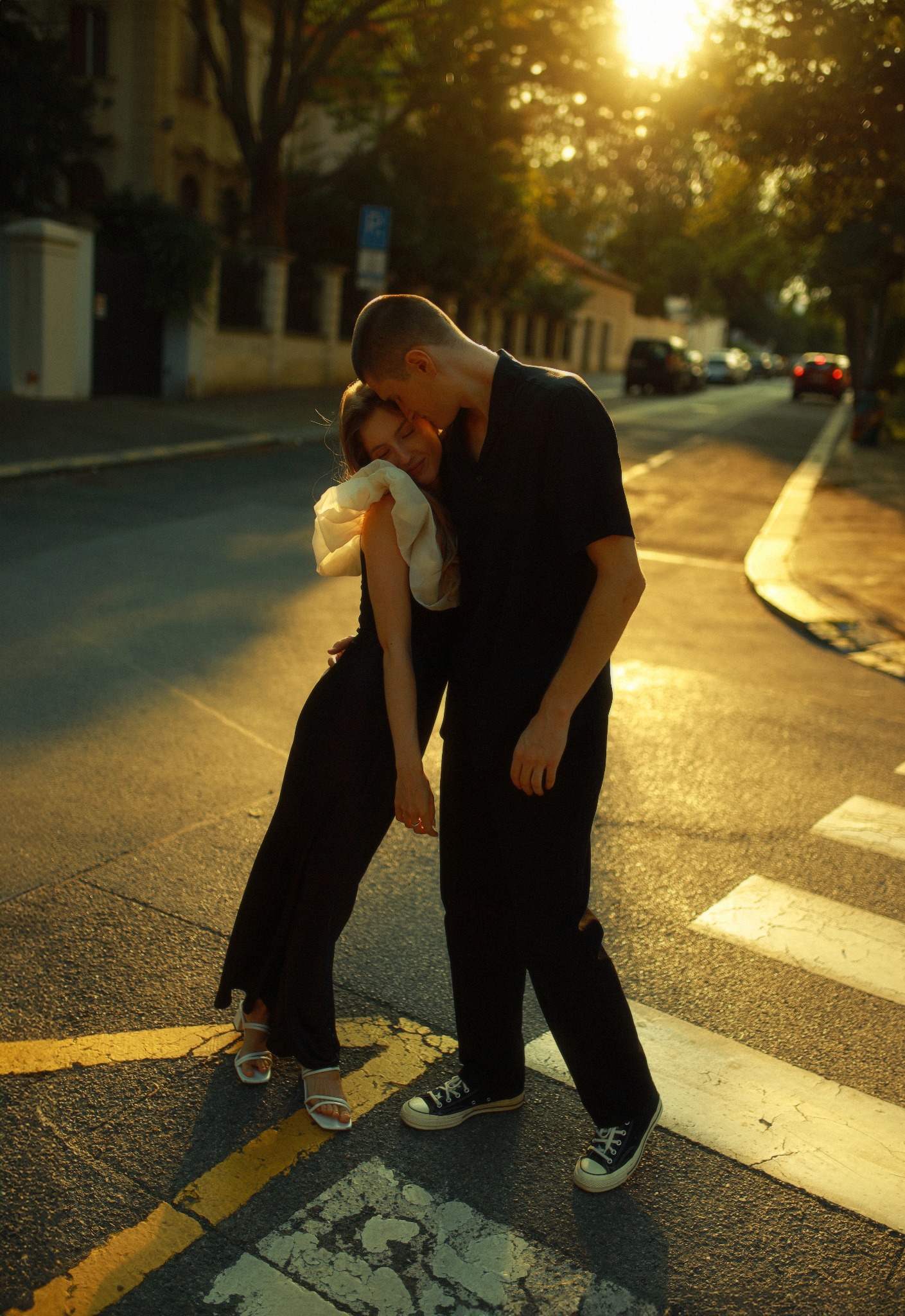 Para w czarnych ubraniach, on całuje ją w głowę, stoją na pasach dla pieszych w ciepłym świetle zachodzącego słońca, ona ma sukienkę z dużym białym elementem na ramieniu.