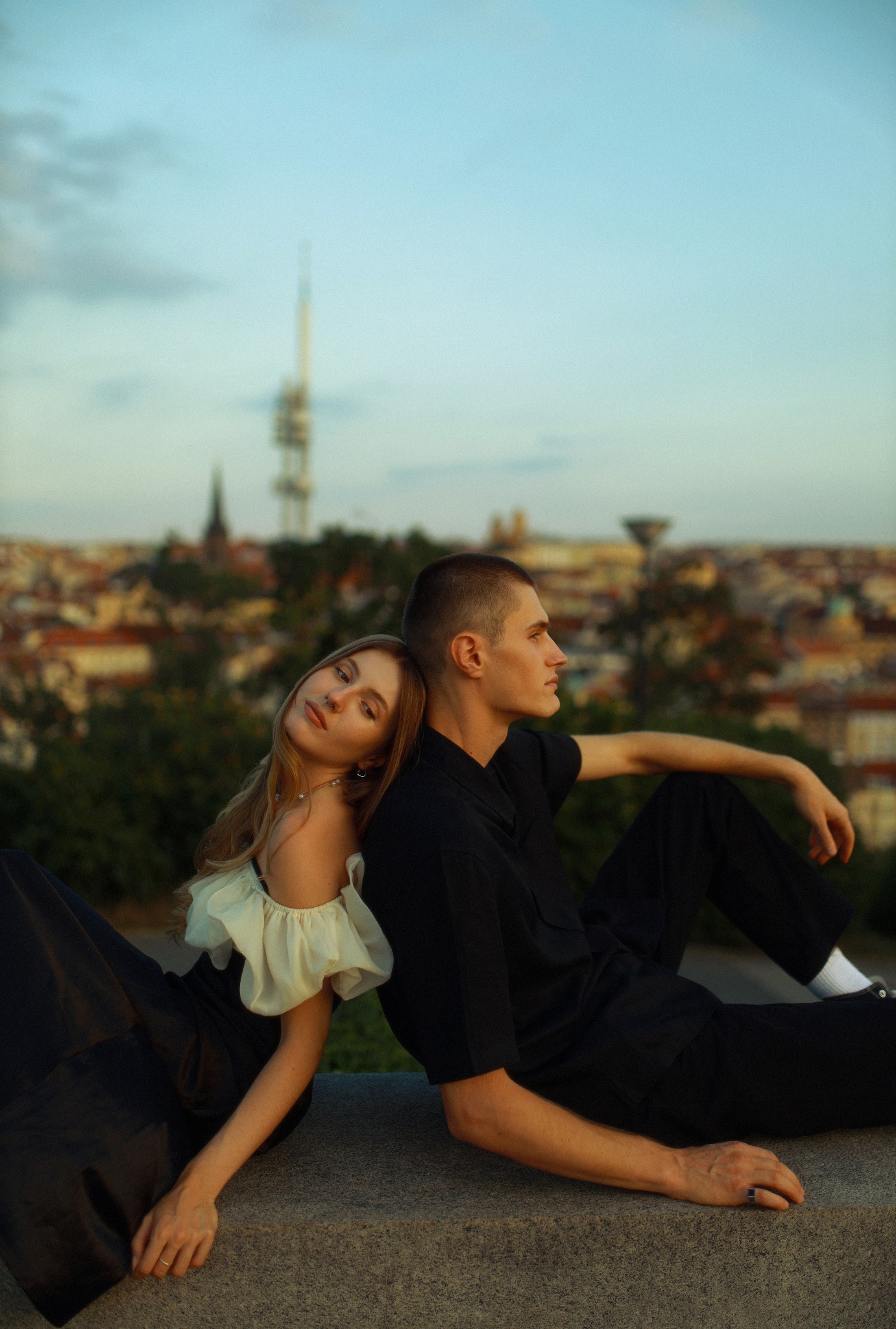 Para siedzi plecami do siebie na tle panoramy Pragi o zachodzie słońca. Kobieta w sukience z falbanami opiera głowę o ramię mężczyzny ubranego na czarno.