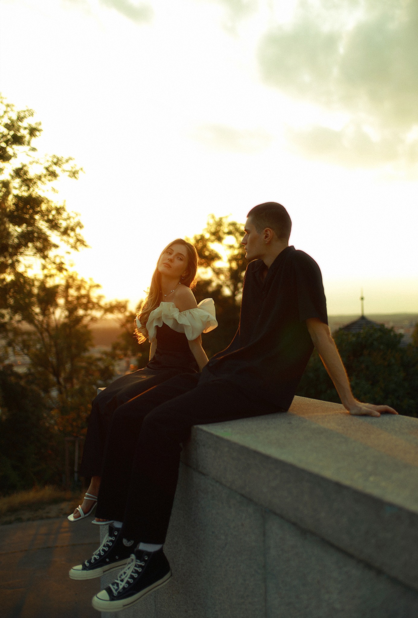 Para siedząca na murku o zachodzie słońca, kobieta w sukience z bufkami i sandałach, mężczyzna w czarnej koszuli i trampkach Converse, w tle panorama miasta.