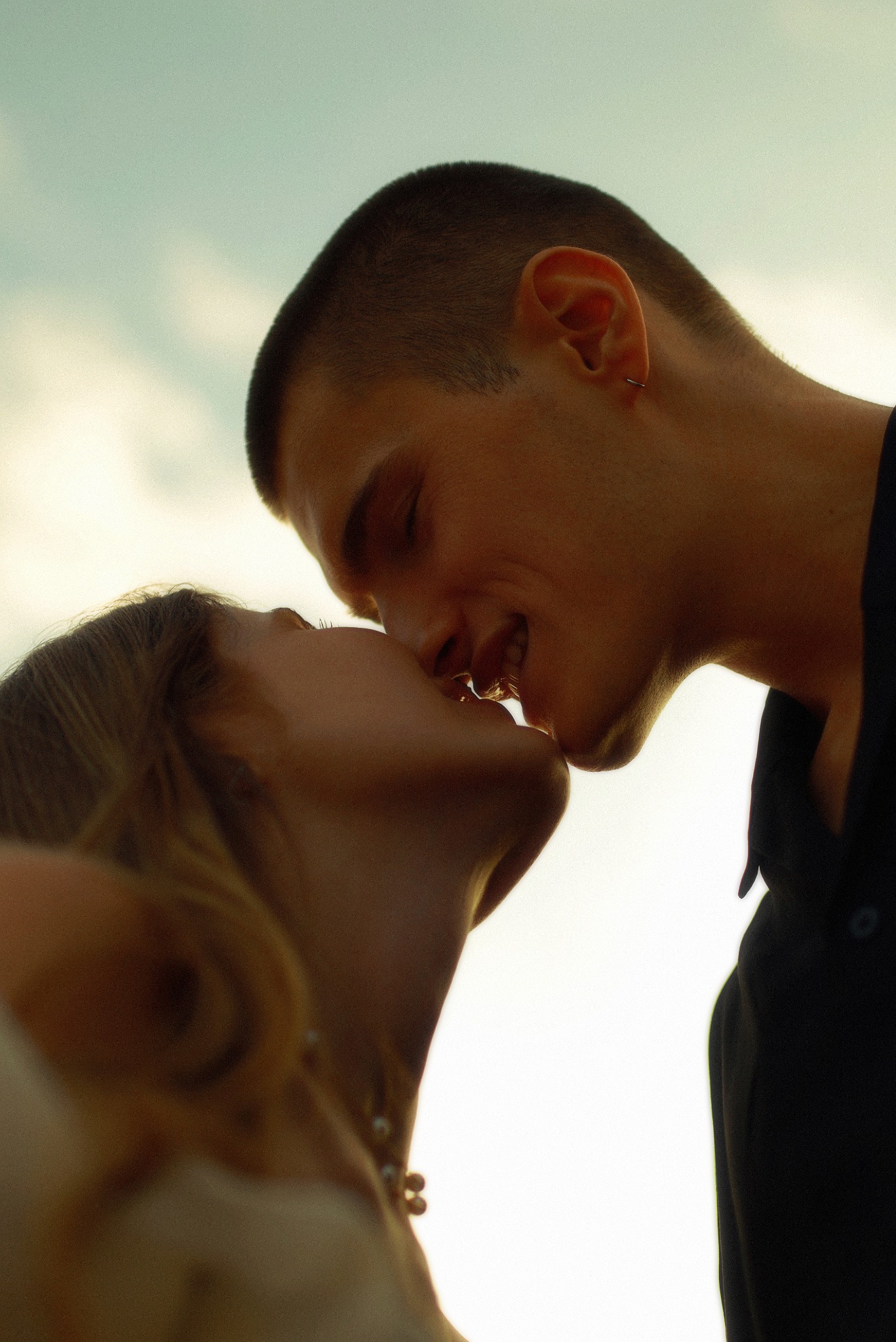 Zbliżenie profilu młodej pary podczas pocałunku, z widocznym kolczykiem w uchu mężczyzny i delikatnym naszyjnikiem kobiety, na tle jasnego nieba.