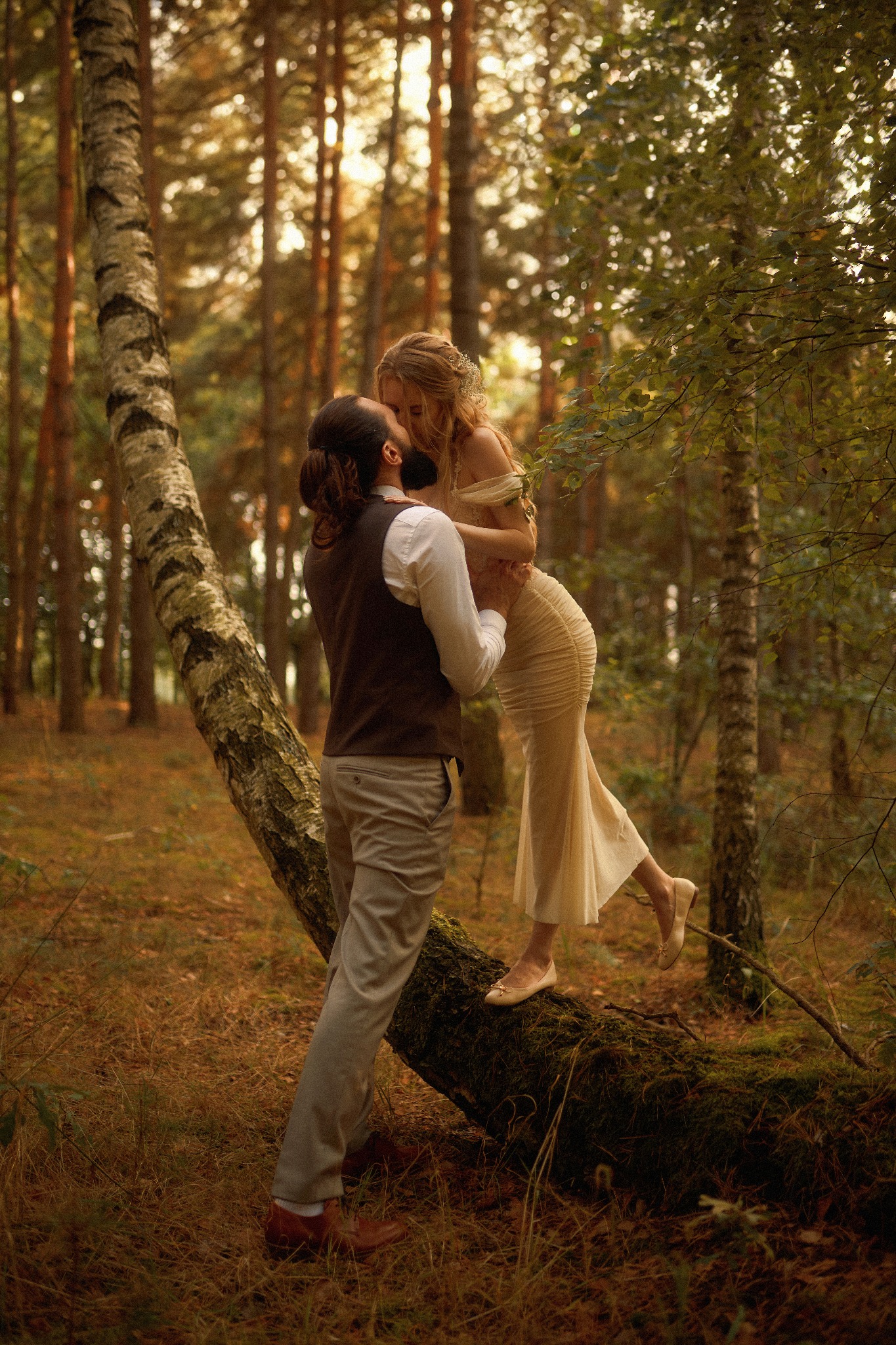 Zakochana para całuje się w lesie; kobieta w długiej kremowej sukni stoi na powalonym pniu drzewa, opierając się o mężczyznę w kamizelce i spodniach w kant.