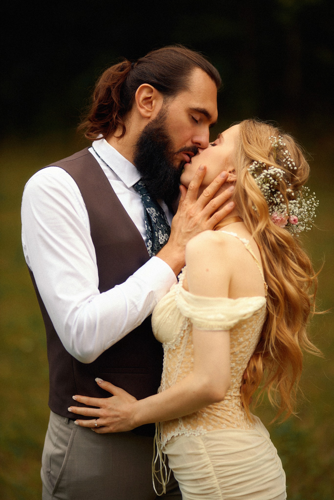 Romantyczny pocałunek pary młodej w stylizowanej sesji ślubnej, panna młoda z kwiatami we włosach, pan młody w kamizelce i wzorzystym krawacie.