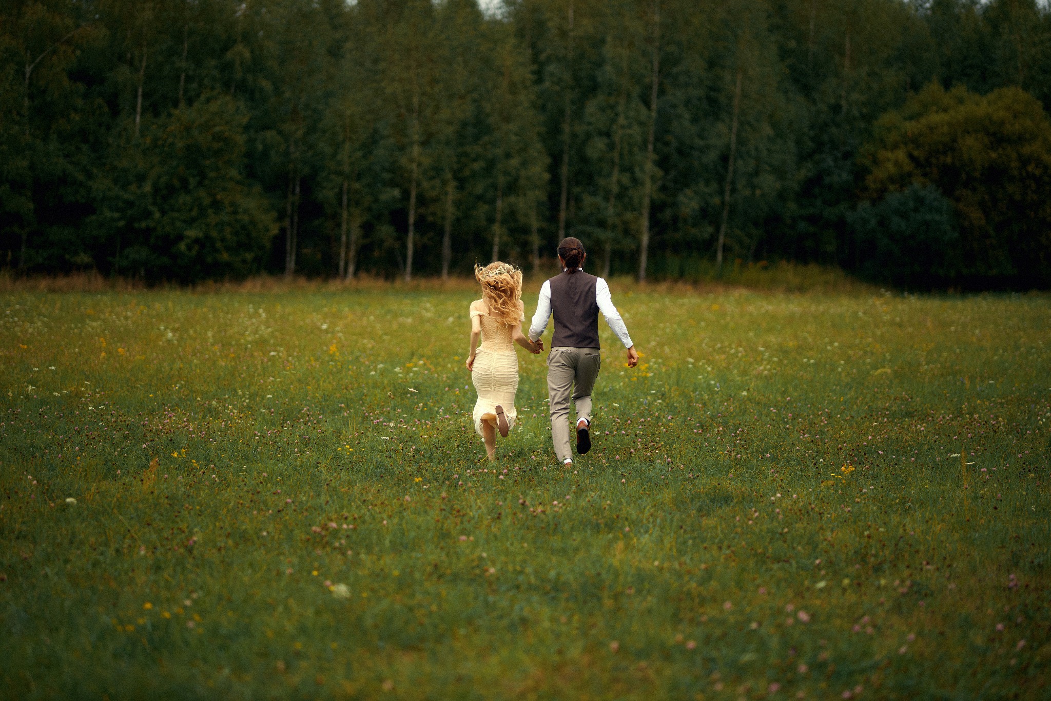 Para młoda biegnie trzymając się za ręce przez łąkę pełną kwiatów w kierunku lasu.