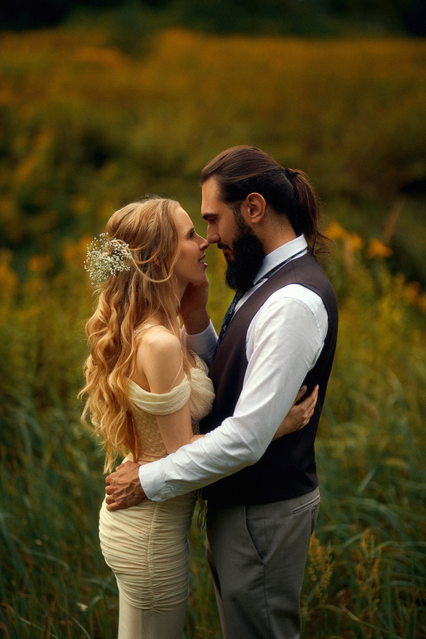 Para narzeczonych w objęciach, panna młoda z kwiatami we włosach, pan młody z brodą i włosami spiętymi w kucyk, na tle łąki z zielono-żółtą roślinnością.