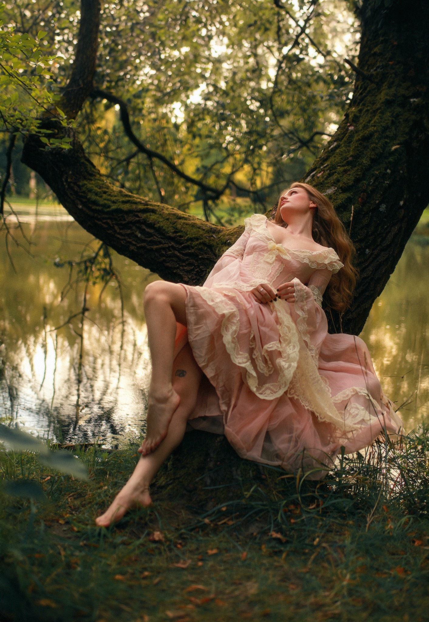 Kobieta w zwiewnej, różowej sukni z koronkowymi zdobieniami siedzi w rozwidleniu starego, omszałego drzewa nad brzegiem jeziora, zamyślona i spoglądająca w górę.