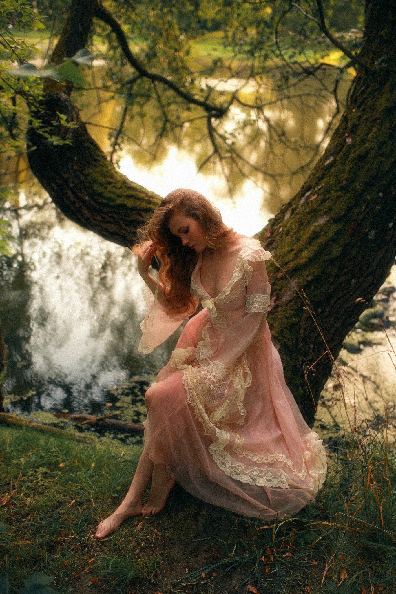 Kobieta w zwiewnej, koronkowej sukni siedzi pod rozłożystym, omszałym drzewem nad brzegiem jeziora; refleksy światła na wodzie, melancholijny nastrój.