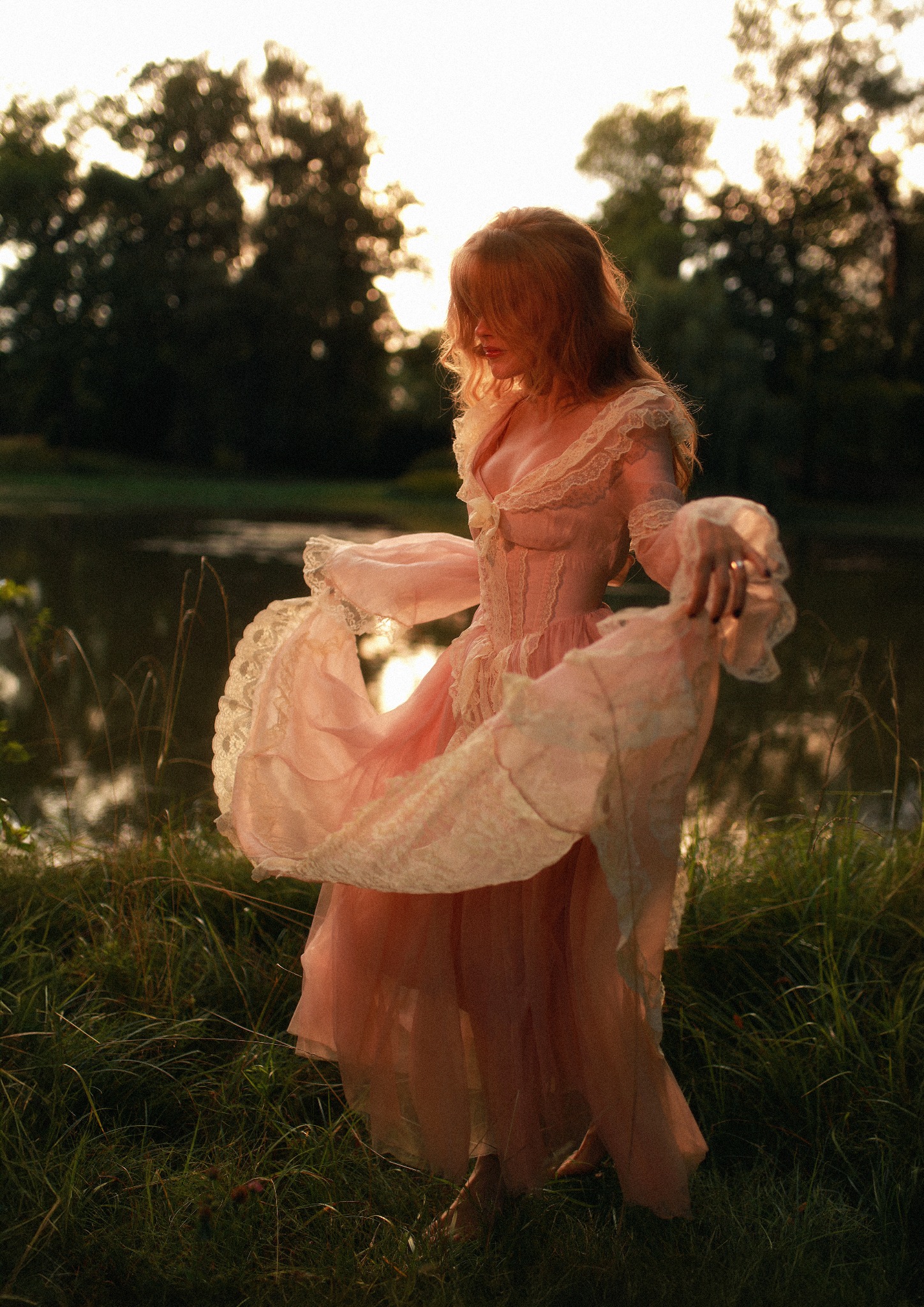 Kobieta w zwiewnej, różowej sukni z koronkowymi zdobieniami, wirująca na tle jeziora i zachodzącego słońca, w otoczeniu wysokiej trawy.