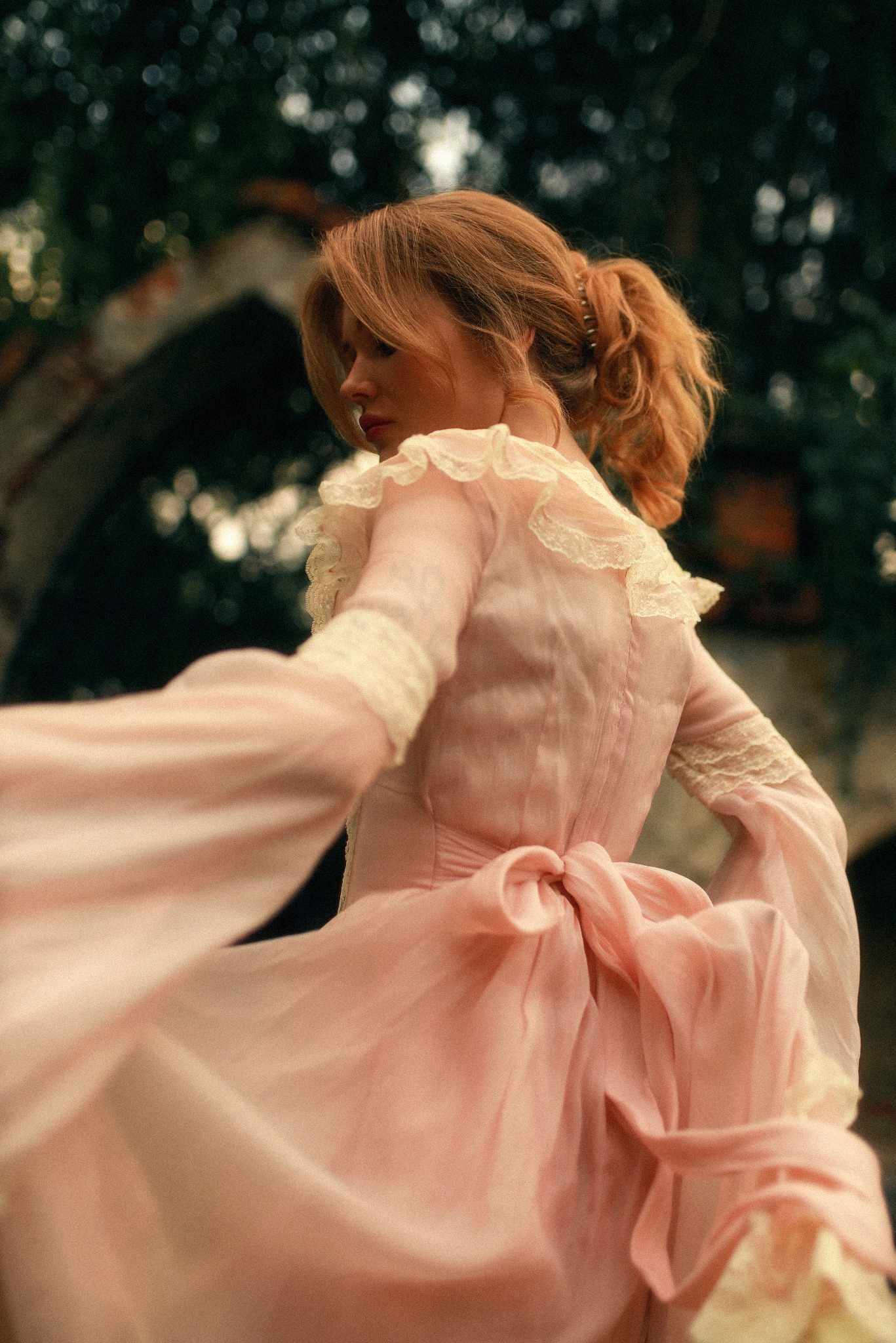 Kobieta z rudymi włosami spiętymi w kucyk, ubrana w zwiewną, różową sukienkę z koronkowymi wstawkami, odwraca się plecami w plenerze.