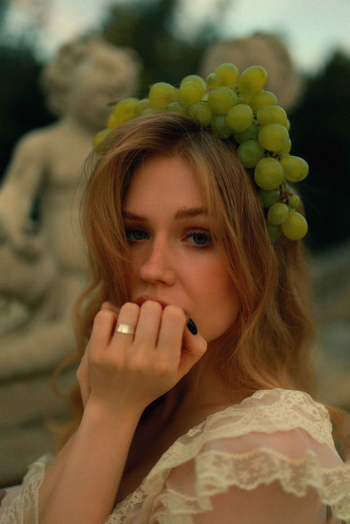 Portret młodej kobiety z wiankiem z zielonych winogron na głowie, w tle rozmyta statua aniołka, dłoń z pierścionkiem przy ustach, delikatna koronka na ramieniu.