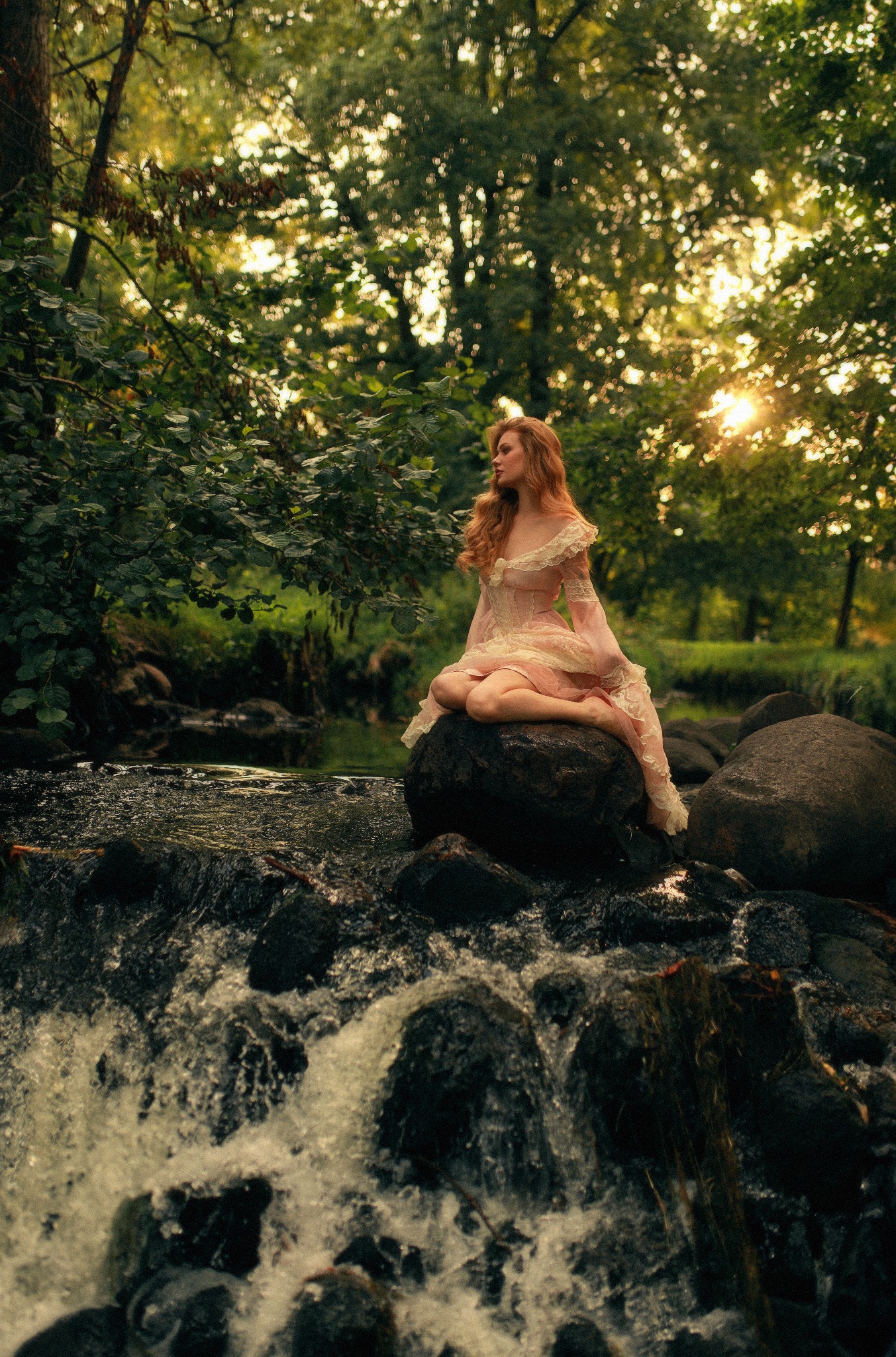 Rudowłosa kobieta w zwiewnej, koronkowej sukni siedzi na kamieniu w strumieniu, otoczona zielenią drzew i promieniami zachodzącego słońca.
