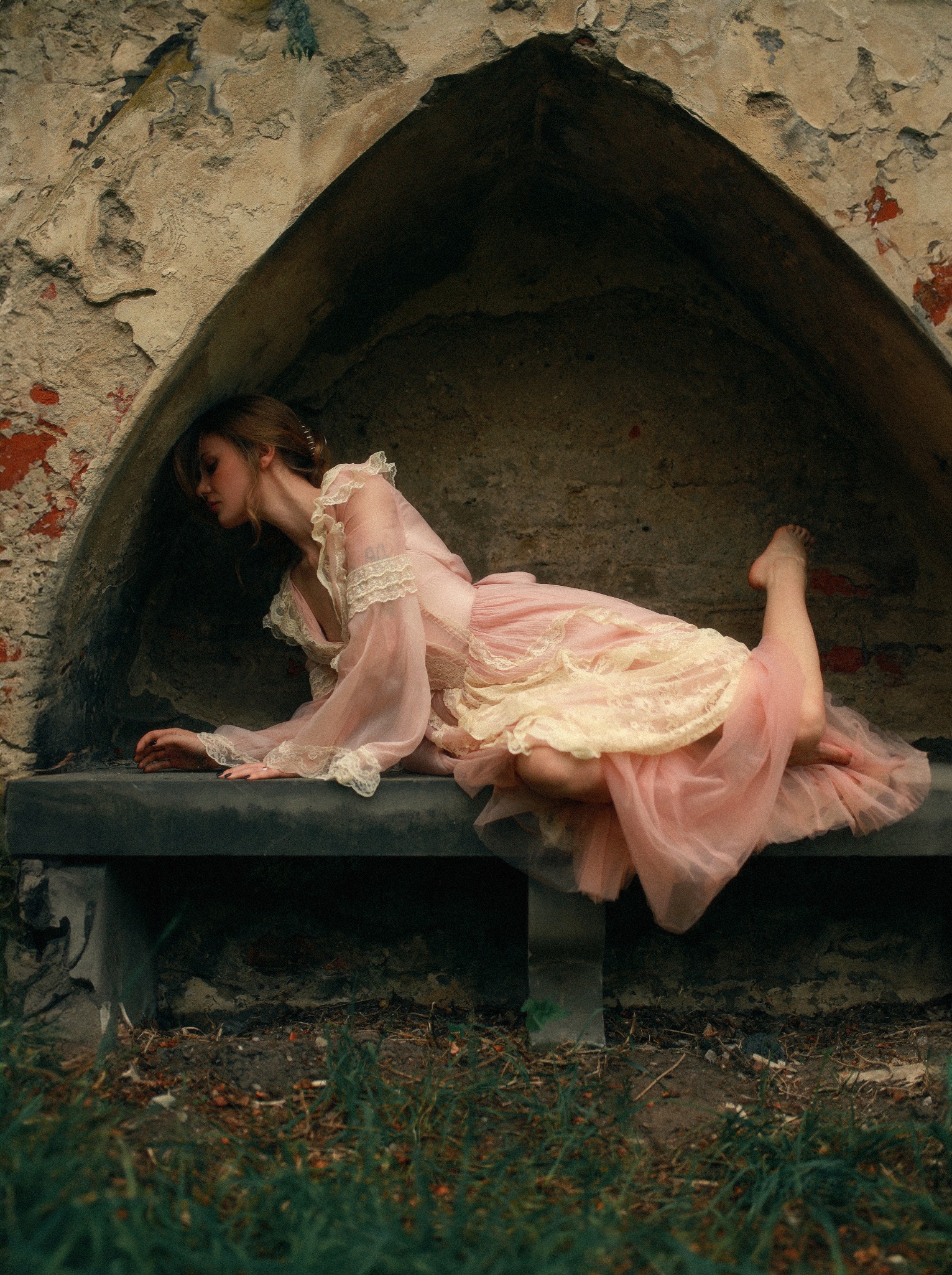 Kobieta w zwiewnej, różowej sukni z koronką leży na kamiennej ławce w niszy muru w Łodzi. Artystyczne ujęcie z trawiastym tłem.