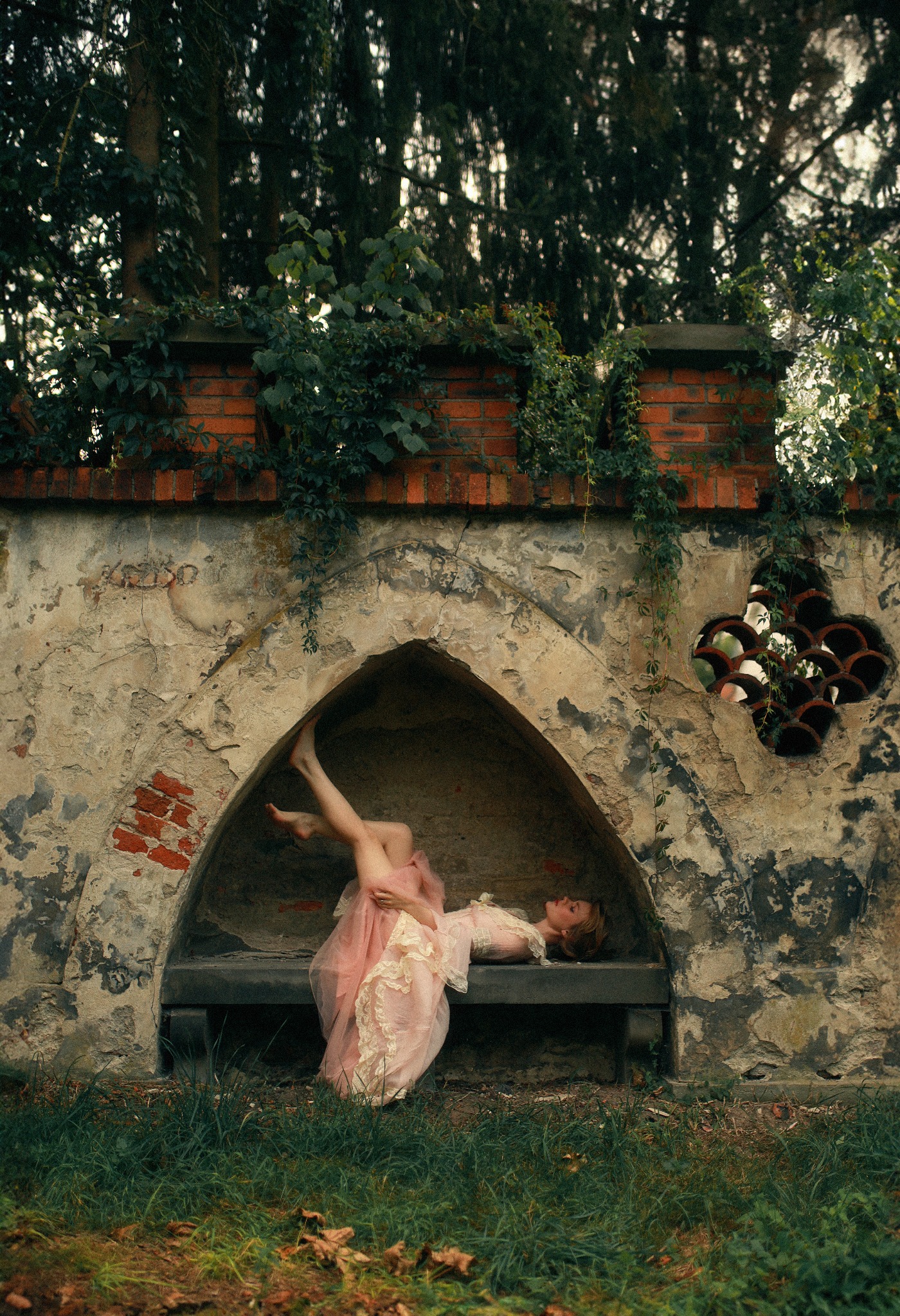 Kobieta w zwiewnej, różowej sukience z koronkowymi detalami leży na kamiennej ławce w niszy starego, ceglanego muru z roślinnością, nogi uniesione do góry, artystyczna sesja w plenerze.