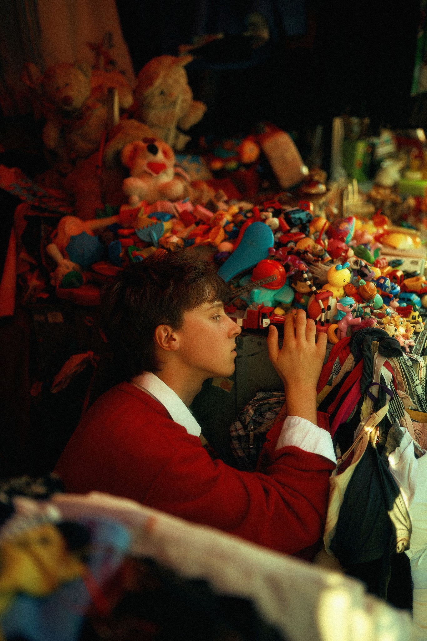 Młoda osoba w czerwonym swetrze ogląda stoisko z zabawkami i ubraniami na pchlim targu, z profilu, skupiona na wyborze.